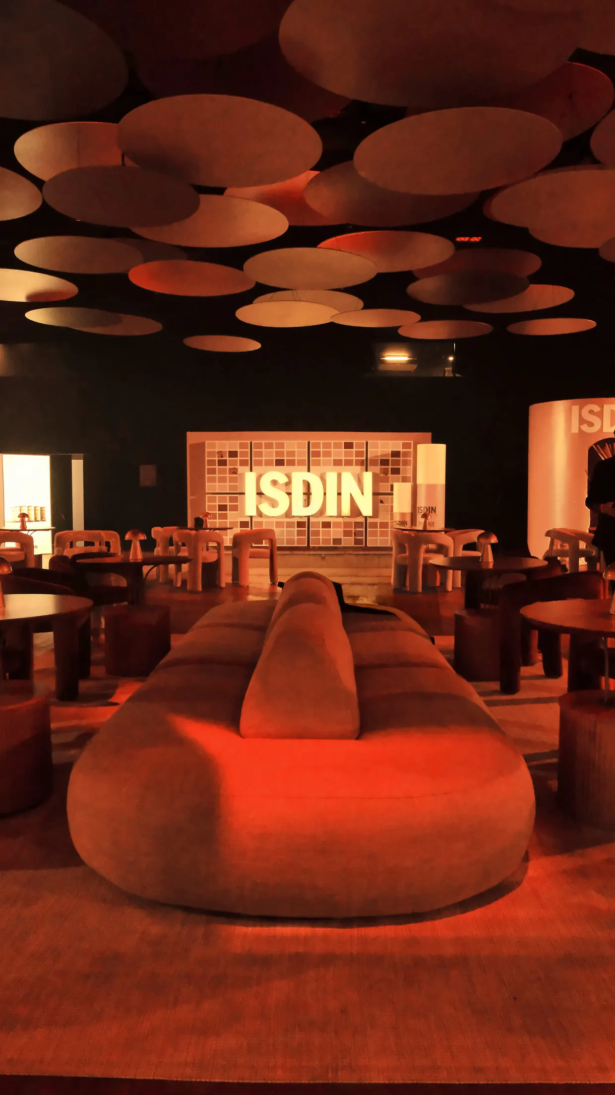 Lounge do evento da ISDIN com sofá curvo, mesas e luminárias circulares no teto, sob luz alaranjada.
