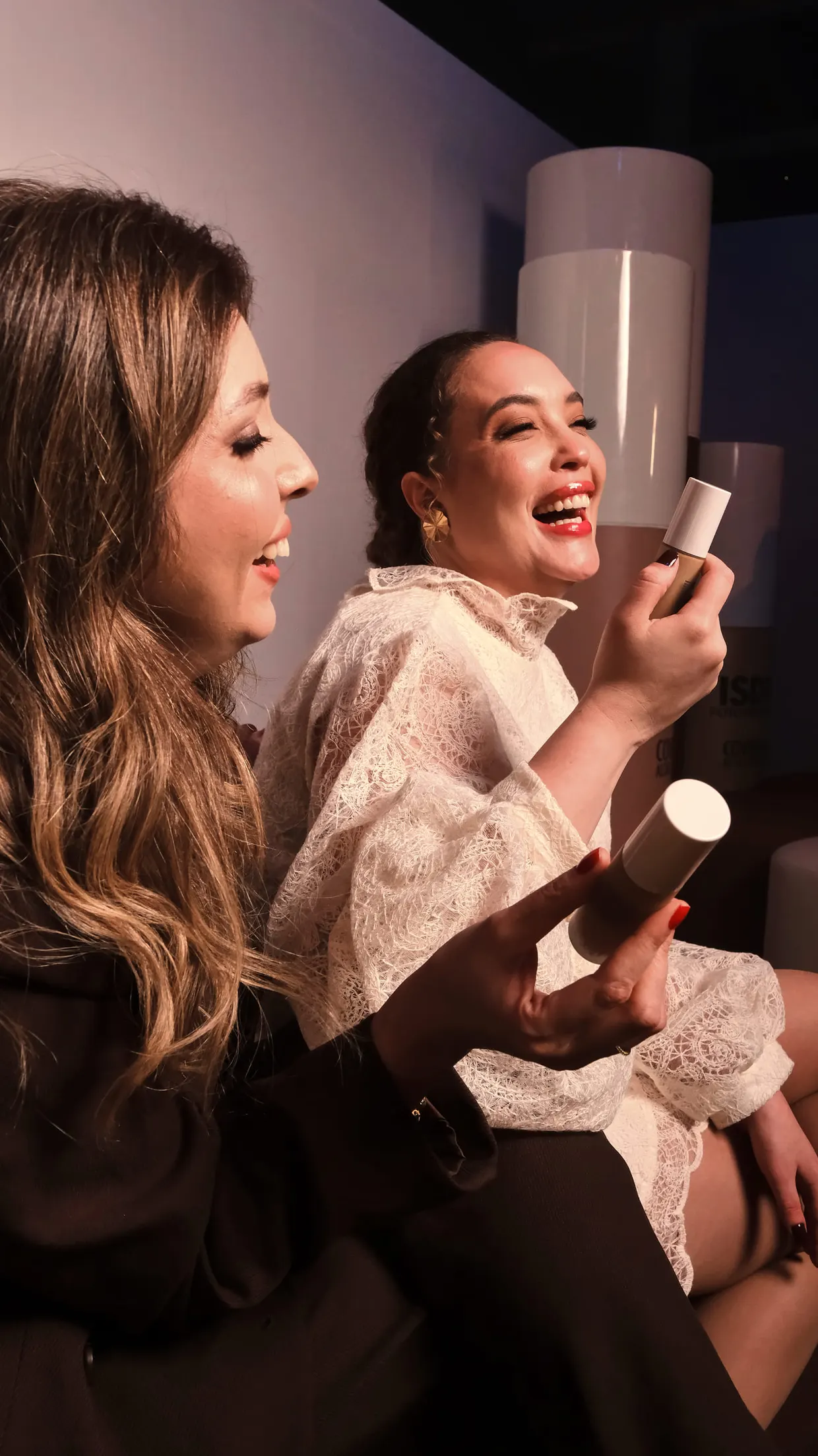 Duas mulheres riem sentadas lado a lado, testando frascos de base.