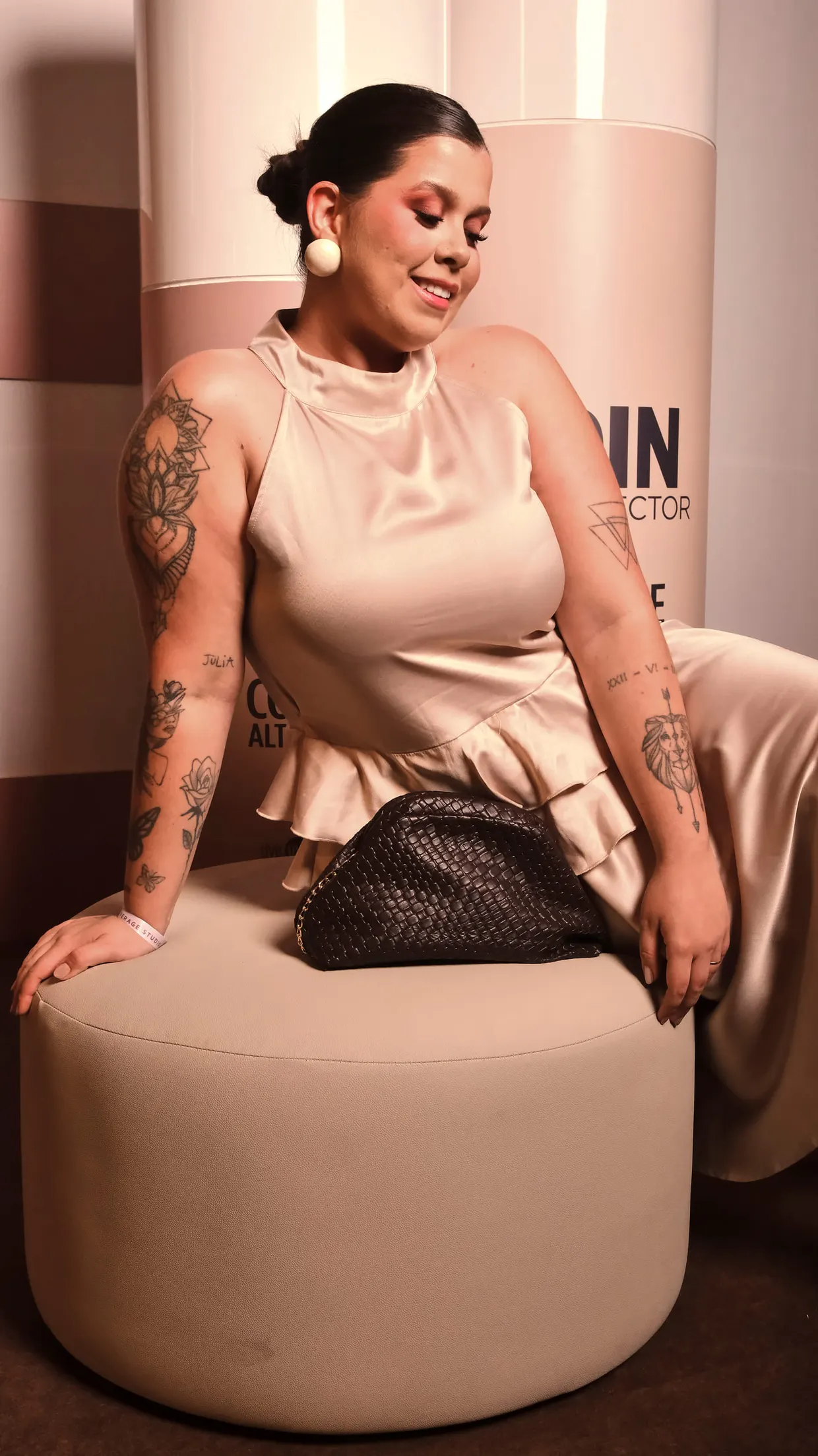 Mulher tatuada de blusa acetinada clara sorri sentada em um pufe, com uma bolsa trançada ao lado.
