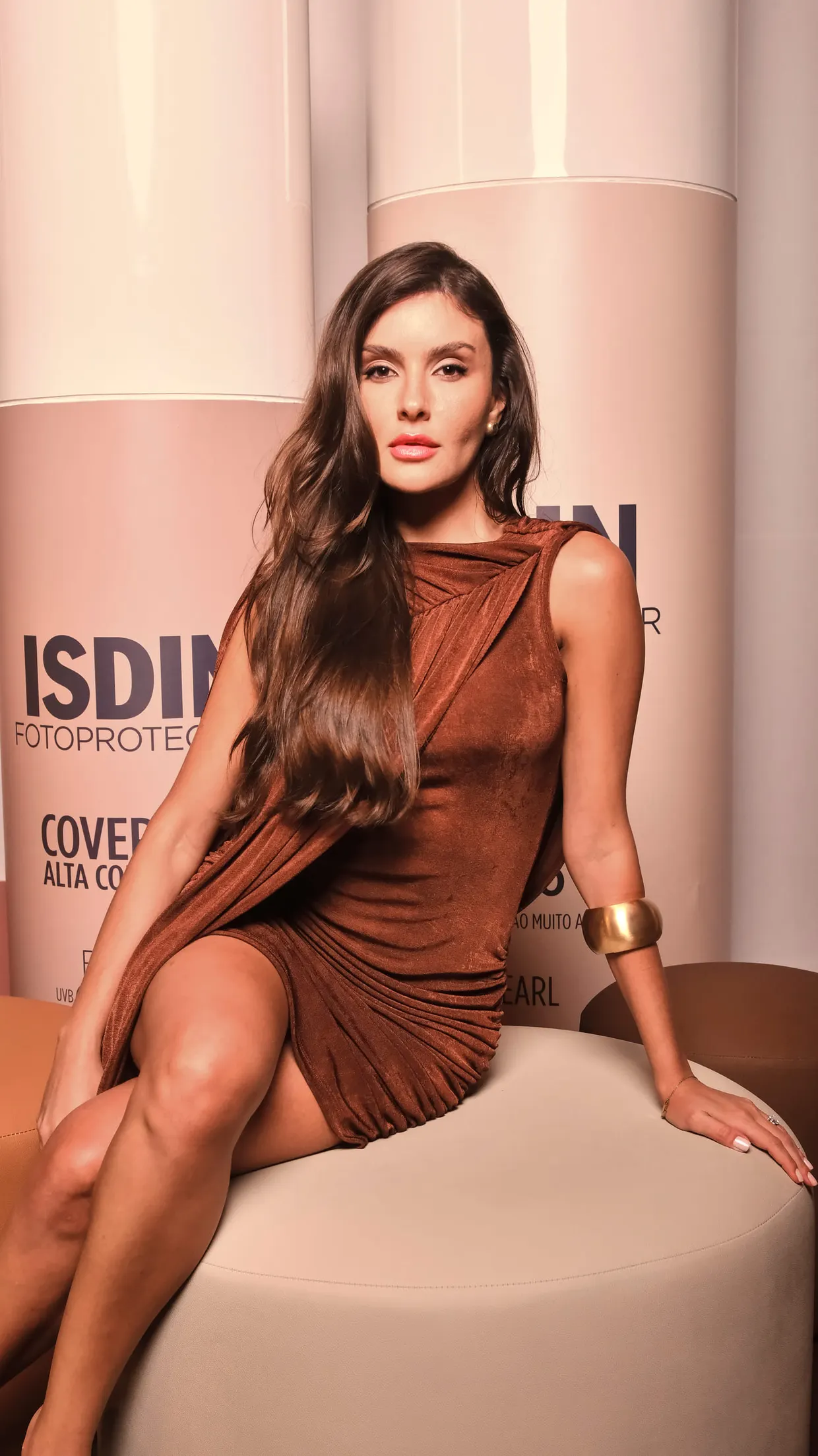 Mulher de cabelos longos e vestido marrom drapeado senta-se em um pufe diante do cenário da ISDIN, com um bracelete dourado.