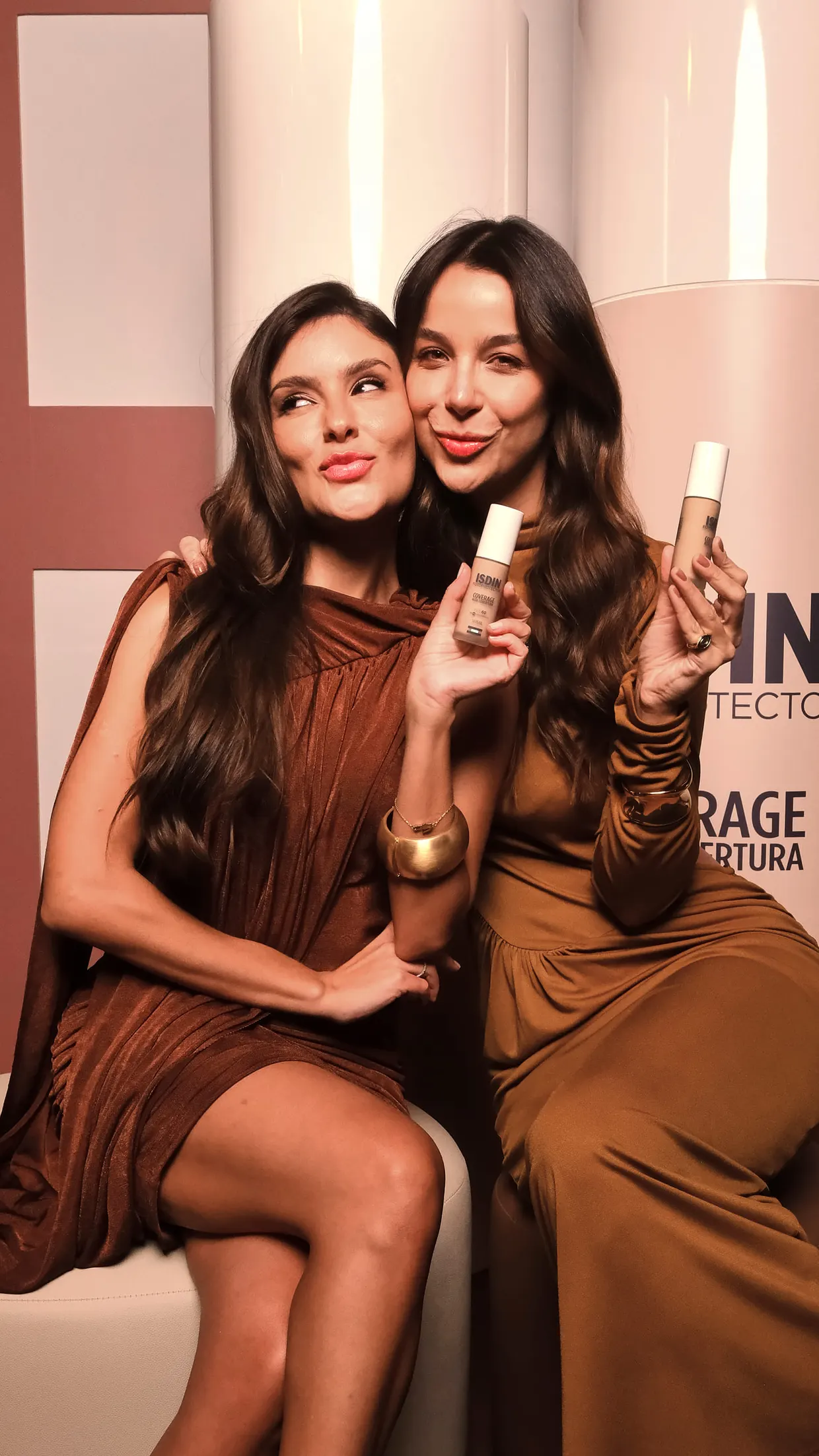 Duas amigas abraçadas fazem biquinho e mostram frascos de base, sentadas diante do cenário.