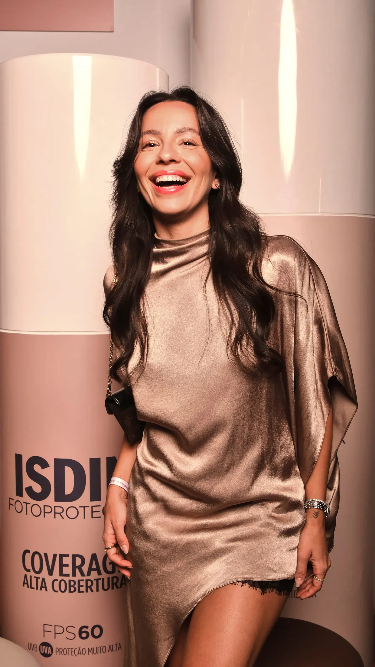 Mulher de vestido acetinado dourado gargalha diante da coluna com a marca ISDIN Fotoprotector.