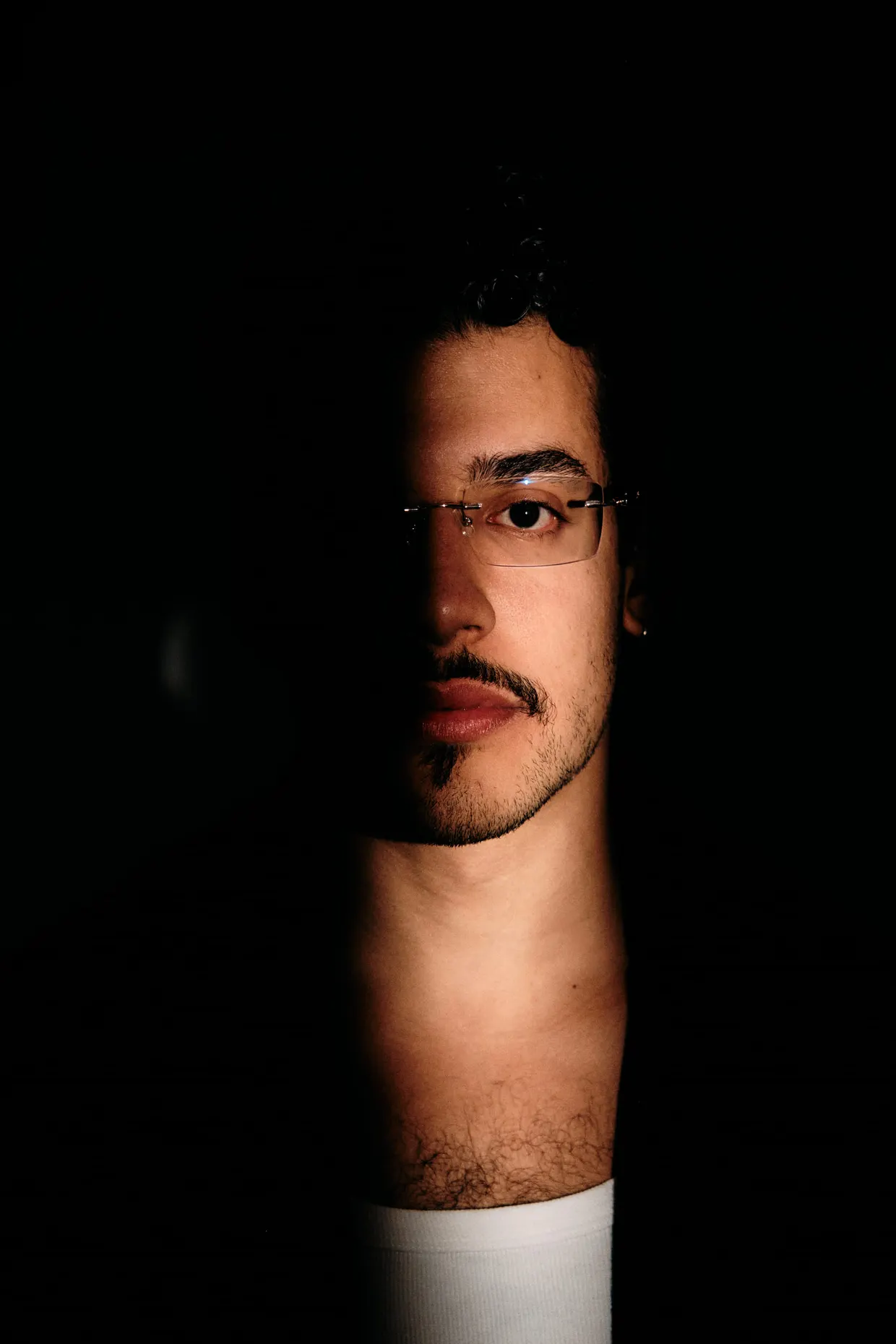 Uma faixa de luz ilumina parte do rosto de um homem de óculos em fundo preto.